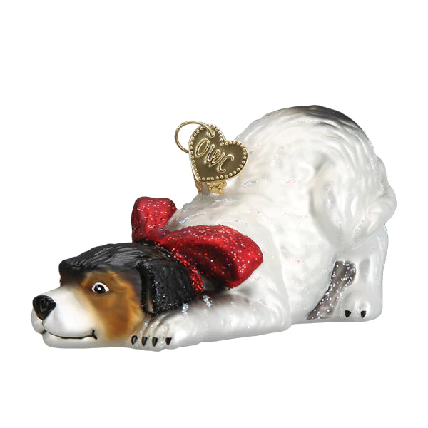 The Norman Rockell Signature Dog Ornament from Old World Christmas (product number 12666) is a festive decoration displaying white and black fur with a red bow around its neck. It depicts the playful stance of the dog, complete with a golden heart-shaped tag on its collar.