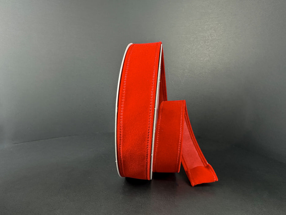 The 7/8 by 10 Yard Red Velvet Ribbon 131805-13 stands upright against a dark background, its bright red velvet and white-edged length unfurled and laying beside the roll.