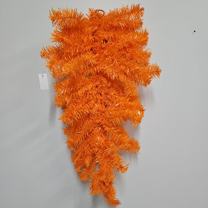 The 24 Halloween Teardrop Orange 82298-OR hangs upside down on a gray wall, featuring bright orange artificial pine branches and a small white tag, creating a striking Halloween decoration.
