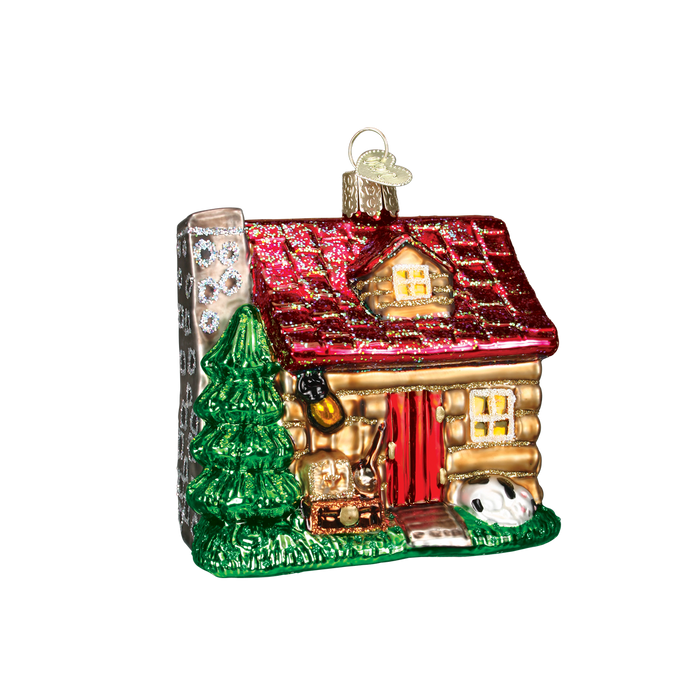 The Lake Cabin Old World Christmas Ornament 20026 is a glass ornament shaped like a cozy lake cabin with a red roof, chimney, pine tree, front door, lamp, treasure chest, and white animal on green grass. Includes hanging loop.