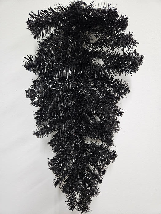 The 24 Halloween Teardrop Black 82298-BK hangs upside down against a white wall, featuring shiny black needle tinsel branches with no decorations.