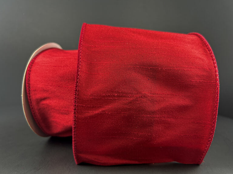 A spool of 4X10Y Red Faux Dupioni Ribbon (72434-04-12), featuring a wide, shiny, slightly textured surface, lies on its side against a plain, dark background.