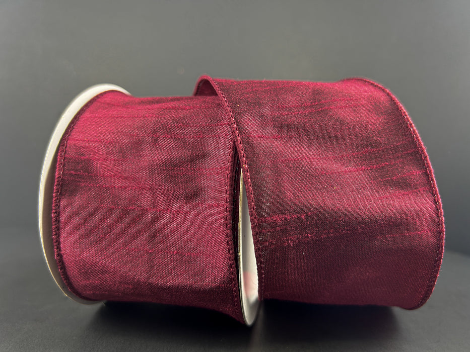 The 2.5"X10Y Burgundy Faux Dupioni Ribbon 72434-40-10, a must-have in craft supplies, is elegantly showcased against a dark backdrop. The ribbon is partially unrolled, highlighting its luxurious texture and sheen.