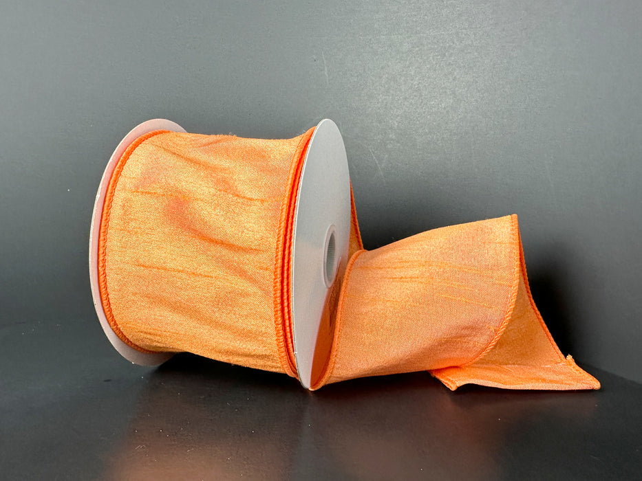 The 2.5X10Y Peach Faux Dupioni Ribbon 72434-40-23, vibrant with a satin-like sheen, is partially unspooled against a dark background, draped loosely to the side on its circular spool.