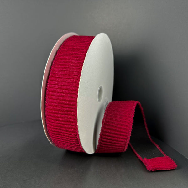 A spool of 1.5x10y Dk Red Ribbed Velvet Ribbon (73561-09-10) stands upright against a dark background, with a length of its deep red ribbon unrolled and lying flat beside it.