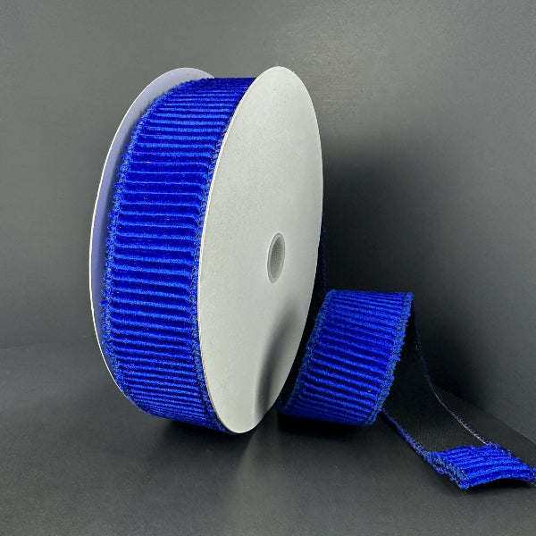 A spool of 1.5X10Y Deep Blue Ribbed Velvet Ribbon (73561-09-25) stands upright against a dark background, with part of the ribbon unrolled and lying flat in front.