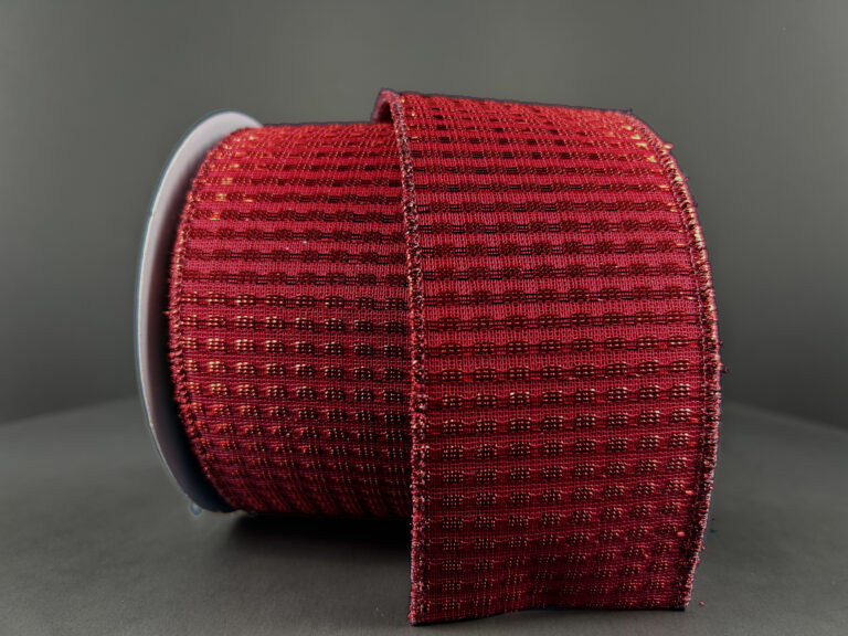 A 2.5X10Y Red Metallic Rishi Solid Ribbon (78496-09-12) with a textured grid-like pattern rests unrolled on a gray surface, showing off its intricate woven design against a matching gray background.