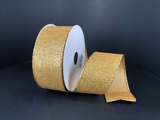 A spool of 1.5X10Y Gold All Flat Glitter X820609-15 ribbon sits on a dark surface, partially unrolled to show its sparkling gold texture and tidy cut edge.