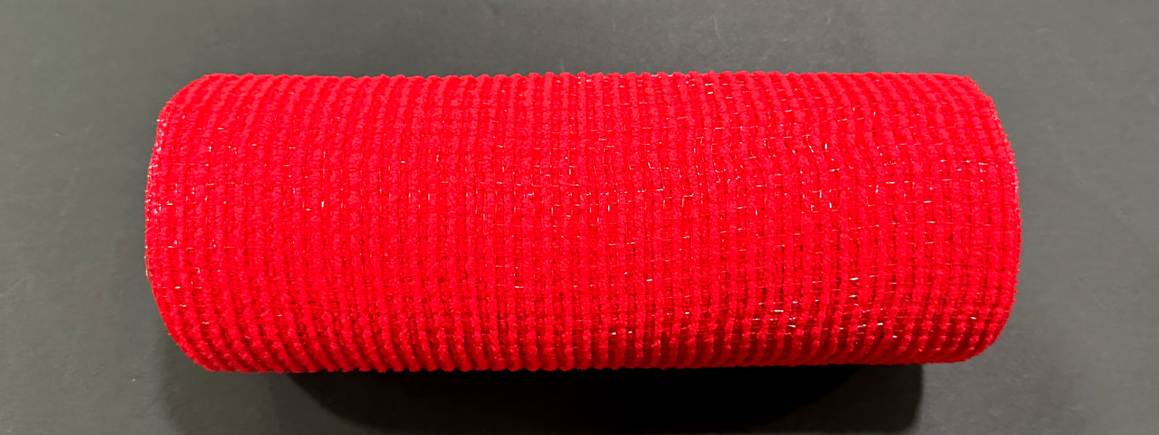 A 10X10Y Red All Snow Fuzz Mesh (XB253610-12) roll rests horizontally on a dark flat surface, displaying its vibrant color and tightly woven, neatly cylindrical form.