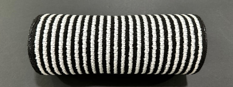 A cylindrical object wrapped in 10X10Y Black-White All Snow Fuzz Stripe Mesh XB253810-20, featuring alternating black and white vertical stripes, is placed on a dark, flat surface.