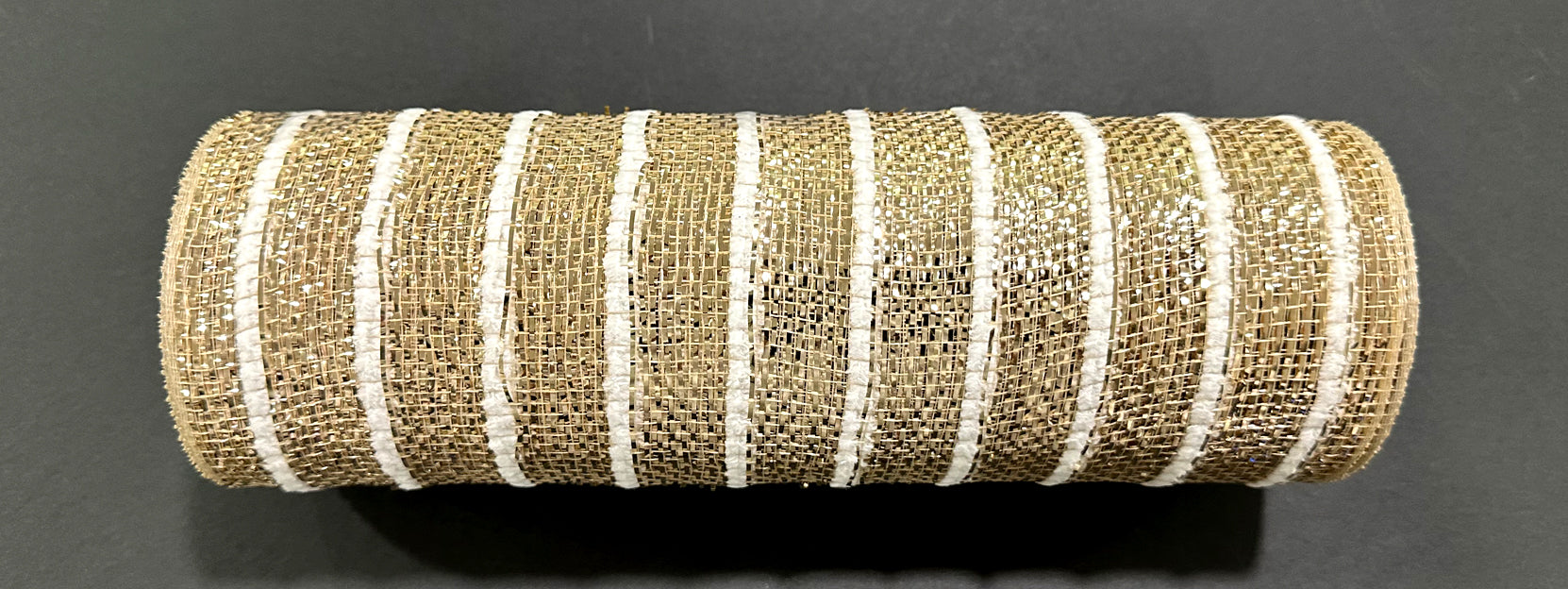 A 10X10Y Champagne Heavy Metallic Snowball Mesh ribbon (XB254610-14) with white stripes lies on a dark surface, its metallic woven texture and cylindrical shape reflecting the light.