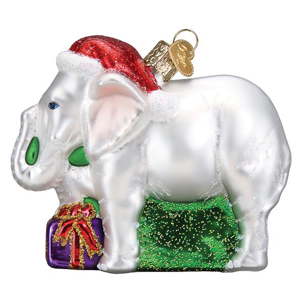 The White Elephant Ornament Old World Christmas Ornament 12592 features a white elephant in a Santa hat, standing on a green glittery base next to a purple gift—perfect for holiday gifting and white elephant exchanges.