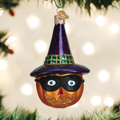 The Masked Witch Jack Olantern Ornament by Old World Christmas (26093), featuring a glittery purple witch hat, green band, black mask, and gold grin, hangs on a Christmas tree with blurred lights in the background.