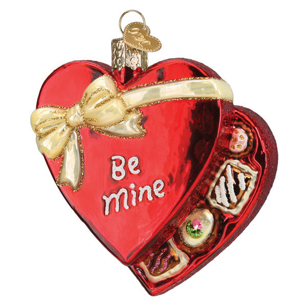 The Valentine Chocolates Ornament by Old World Christmas (32540) is a red, heart-shaped decoration with a gold bow and "Be mine" text, featuring an open lid that reveals assorted chocolates—an ideal Valentine's Day gift.