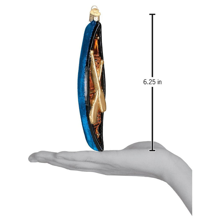 A hand holds the Canoe 46033 Old World Christmas Ornament Assorted, a 6.25-inch tall hand-painted blue and gold glass ornament featuring two crossed oars, as shown by the vertical measuring line.