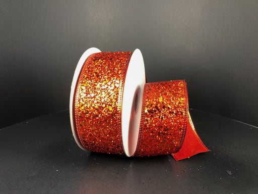 Two rolls of Orange Super Glitter 1.5 x 10Y Ribbon (75102-09-19) with white spools rest on a dark surface against a black background; one stands upright while the other lies on its side, partially unrolled and sparkling.