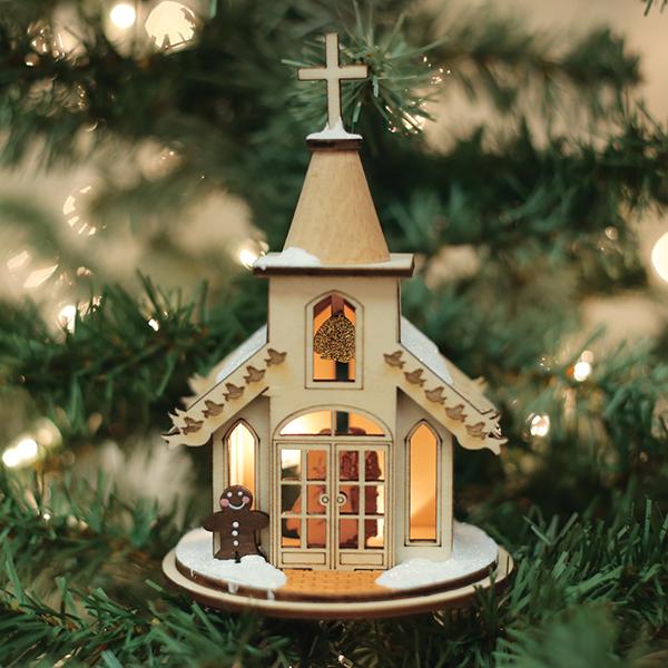 The Wedding Chapel Old World Christmas Ornament 80002, featuring a lit church with snowy rooftops, a wreath, and a gingerbread figure, hangs on the Christmas tree surrounded by glowing lights and green pine needles.