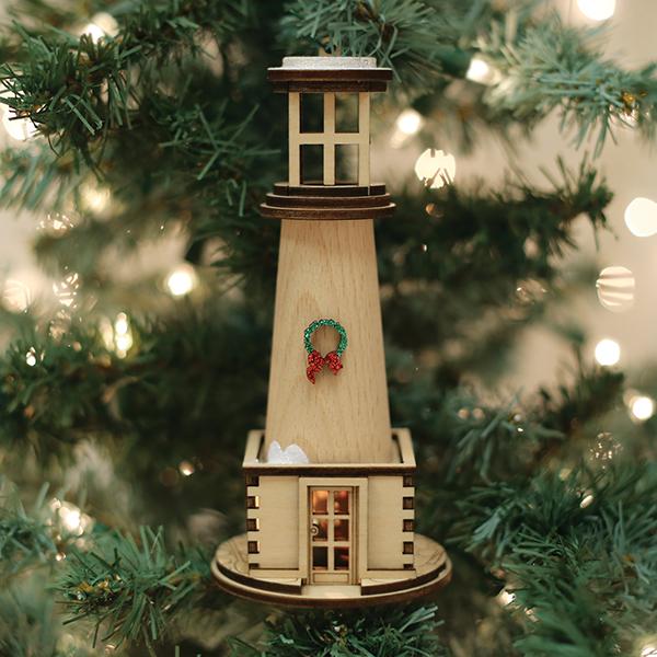 The Holiday Lighthouse Old World Christmas Ornament 80017 hangs on a tree, featuring a small wreath and surrounded by green branches and warm, blurred holiday lights in the background.
