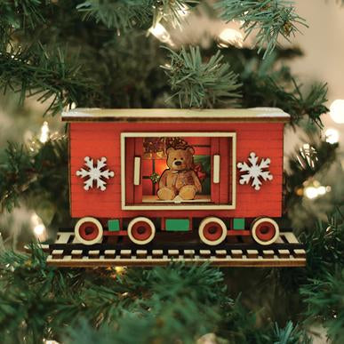 The Santa's North Pole Express Box Car GC139 Old World Christmas Ornament 80036, featuring red color, snowflakes, and a teddy bear inside, hangs on a tree with green branches and white lights.