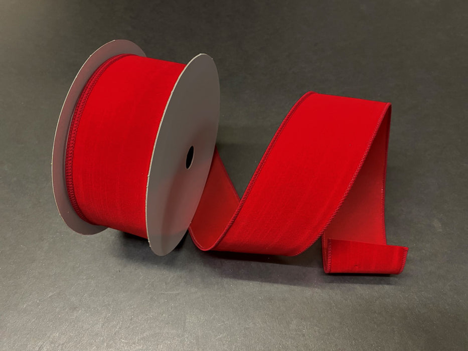 The Christmas Red Velvet 2.5X25Y Ribbon X501140-13B, a vibrant red reminiscent of festive decor, lies partially unrolled on a dark gray surface. This wide wired ribbon adds texture and elegance, perfect for holiday crafts or gift wrapping.