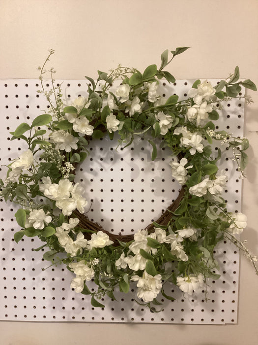 The 22 Cherry Blossom Wreath 62811, embellished with white flowers and green leaves, gracefully hangs on a white pegboard, enhancing any space with its floral charm.