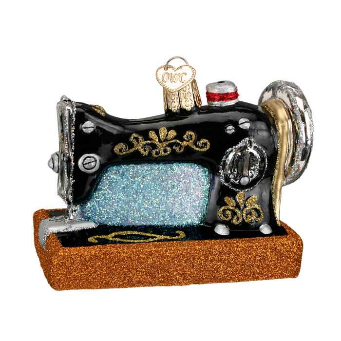 The Sewing Machine 32103 Old World Christmas Ornament is a hand-painted glass ornament shaped like a vintage black sewing machine with gold, silver, glittery blue details, and a brown glittery base.