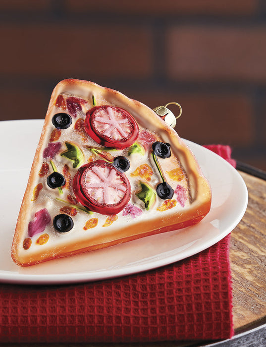 The RAZ Pizza Slice Christmas Ornament, featuring colorful toppings, is displayed on a white plate atop a red textured cloth, with a blurred brick wall in the background.