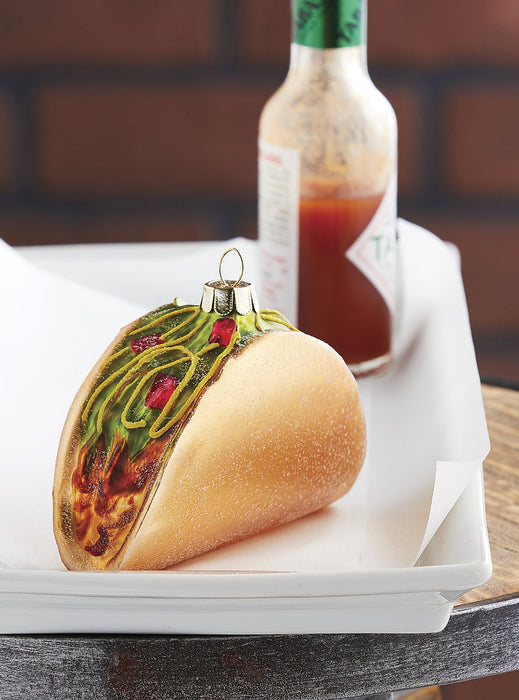 The RAZ Taco Christmas Ornament from the Eat Drink & Be Merry Collection rests on a parchment-lined white tray, while a bottle of hot sauce appears slightly blurred in the background.