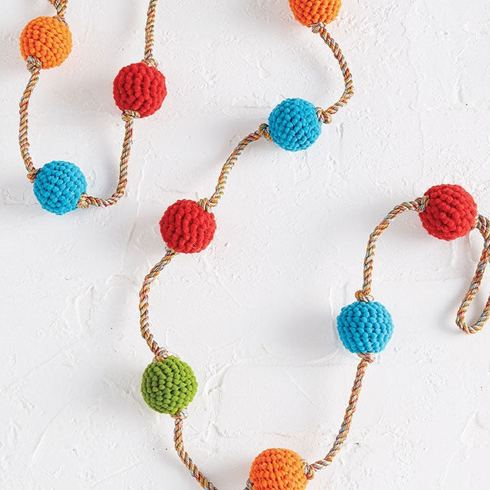 The RAZ Knitted Ball Garland showcases large crochet-covered beads in vibrant orange, red, blue, and green, strung on a twisted multicolored cord and displayed on a textured white surface for a festive touch.