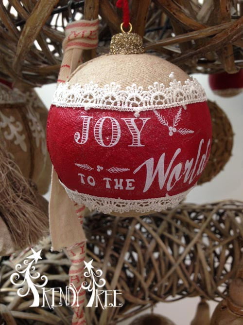 The 4" Joy to the World Ball Ornament Natural Red features red fabric with white lace trim and “Joy to the World” in white, hanging on a rustic tree.
