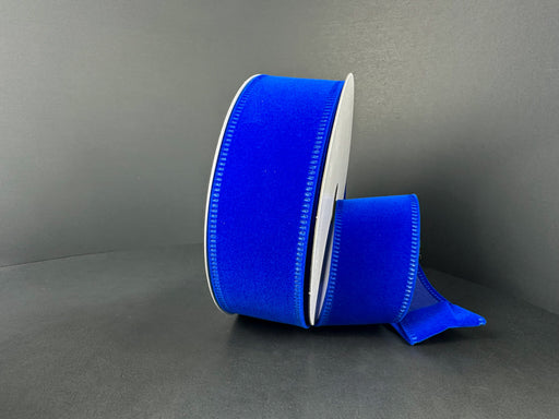 A roll of 1.5 by 10 Yard Royal Blue Velvet Ribbon (131809-39), featuring stitched wired edges, is partially unspooled and shown against a dark background.