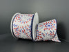 A spool of White Satin/Red-Blue-Silver Glitter Fireworks ribbon (2.5X10Y 42404-40-27) is displayed against a dark background, with its partially unrolled festive design and glitter fireworks motif exuding a celebratory charm.