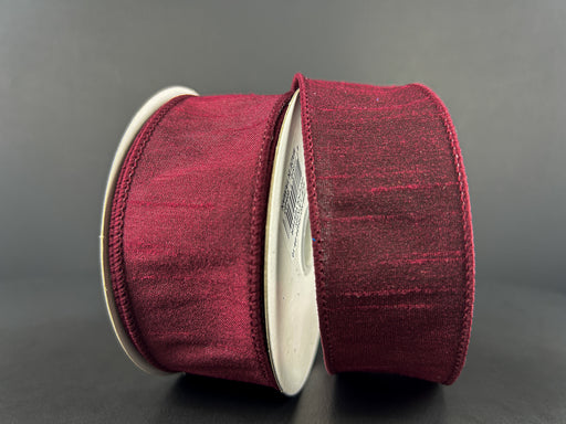 Two spools of 1.5X10Y Burgundy Faux Dupioni Ribbon (72434-09-10) with a subtle sheen are vertically positioned against a dark background. Each spool displays the ribbon wound around a sturdy cardboard center with neatly finished edges.