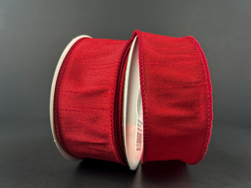 Two rolls of 1.5X10Y Red Faux Dupioni Ribbon 72434-09-12 with a slightly shiny texture stand upright against a dark background, partially unspooled and facing each other.