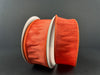 Two large spools of 1.5X10Y Orange Faux Dupioni Ribbon 72434-09-19 are set against a dark background, showcasing their textured appearance and shiny finish, with visible edges that suggest a satin-like or organza material.
