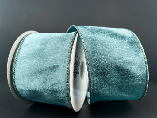 Two spools of 2.5X10Y Dusty Blue Faux Dupioni Ribbon (72434-40-05) sit against a dark background, showcasing their subtle sheen and soft texture.