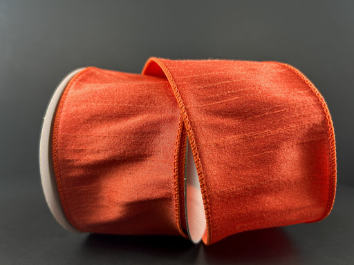 A spool of 2.5X10Y Orange Faux Dupioni Ribbon 72434-40-19 rests on its side against a dark background, with some ribbon unwound and gracefully draped across the spool.