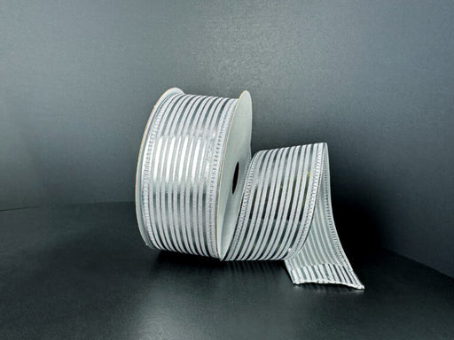 A spool of 1.5X10Y White Satin/Silver Metallic Embossed Vertical Grain Ribbon (76555-09-51) stands upright against a dark background, with a length unrolled and elegantly draped beside it.