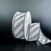 A spool of 1.5X10Y Silver Flat Glitter/White Diagonal Stripes Ribbon (76579-09-01) rests on a dark surface with some ribbon unrolled, showing its shimmering silver and white pattern against a black background.