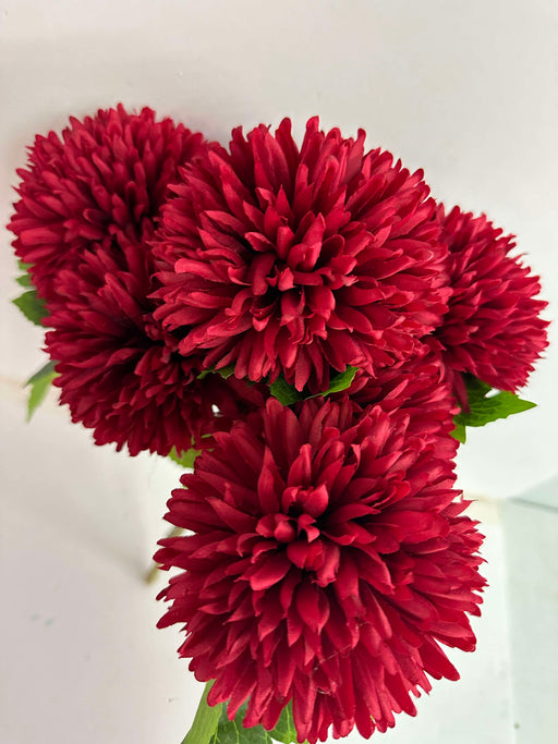 The 16-inch Mum Bush X 7 (84700-Rd) features vibrant red flowers with lush, layered petals. Against a plain white background, they highlight rich color and texture, while green leaves peek through for contrast.