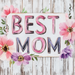 The 10 x 10 Vintage Best Mom Sign TTE-220 features pink and purple text, colorful flowers, and greenery on a rustic white wooden background—a charming décor piece to celebrate moms.
