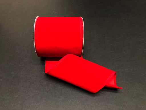A spool of Bright Red Velvet, 4X10Y Ribbon (131804-13) is partially unrolled on a dark surface, displaying its smooth texture and synthetic wired edges as it curves gently.