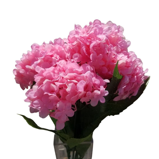 The 19 Pink Hydrangea Bush x 5 Stems resembles a bouquet in full bloom with vibrant pink hydrangeas in a clear glass vase. Clustered with lush green leaves against a white background, it captures natures charm in near-perfect form.