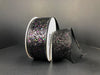 A spool of Black Super Glitter, 1.5X10Y Ribbon (75102-09-21) rests on a dark surface, with some ribbon unrolled to display its sparkling multicolored glitter and shiny, textured finish.