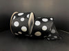 A partially unwound reel of Black Satin/White Glitter Big Dots ribbon, measuring 2.5x10Y (75104-40-21), sits against a dark background, showcasing its synthetic sheen and evenly spaced large white polka dots for striking contrast.
