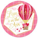 The 12Dia Metal/Foil Love Is In The Air Pink/Red/Hot Pink MD1073 sign features a round illustration of a red and pink heart-adorned hot air balloon beside the cursive Love Is In The Air in glittery gold. Small hearts float below on a soft pink plaid background.