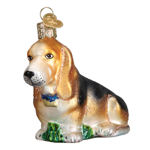 The Basset Hound Old World Christmas Ornament 12473 is a beautifully crafted glass piece in the shape of a basset hound sitting on lush green grass. It features intricate coloring and has a small gold cap at the top, making it perfect for hanging.