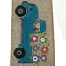 The 2.5 Tan Linen Blue Truck Flowers Ribbon (Q921740-14) features a blue vintage sewing machine with black wheels, colorful flowers at the base, stitched edges, a synthetic wire edge, and comes in a 10-yard length.