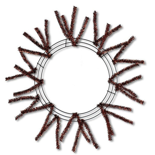 The 24 Chocolate Metallic Pencil Wreath (XX751140) features a circular metal wire frame with dark brown artificial pine branches that radiate outward, creating a sunburst effect reminiscent of holiday designs on a white background.