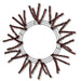 The 24 Chocolate Metallic Pencil Wreath (XX751140) features a circular metal wire frame with dark brown artificial pine branches that radiate outward, creating a sunburst effect reminiscent of holiday designs on a white background.